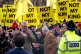 Charles Windsor walks past demonstrators holding signs that read 