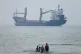A group of people stands in shallow water as a cargo ship appears anchored in the Strait of Hormuz off Bandar Abbas, Iran, June 30, 2026