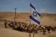 Palestinian shepherd Thiab Draghme leads his flock of sheep and goats past an Israeli flag placed on a road in the northern Jordan Valley of the occupied West Bank, June 23, 2026